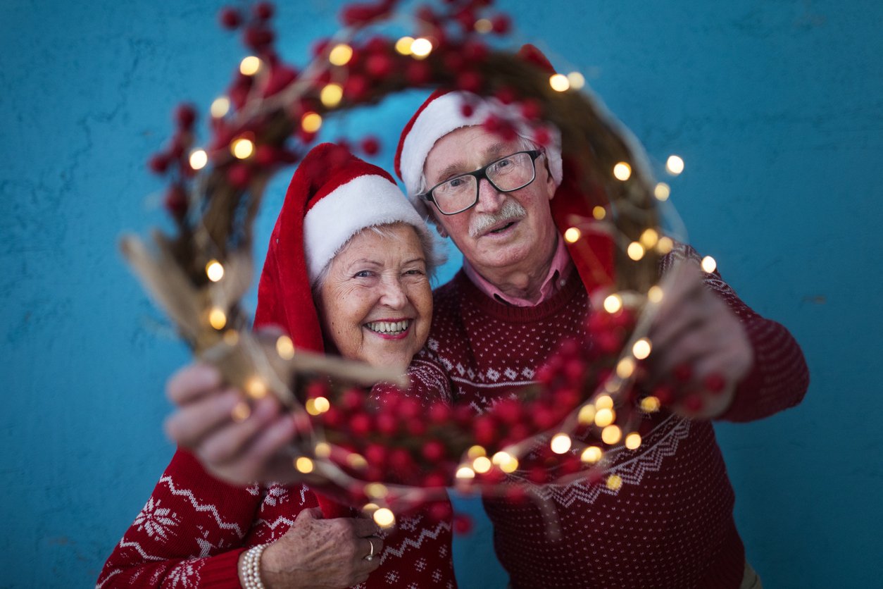 Two elderly people in Santa hats and red sweaters smile while holding a festive wreath with red berries and lights, creating a scene that feels like home in their senior living apartment against a blue background.
