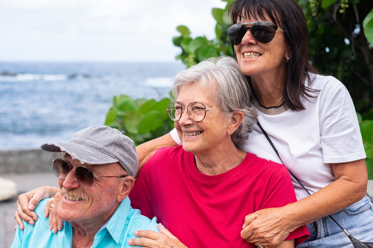 Three older adults, two women and one man, smile while posing together outdoors by the water. All are wearing sunglasses, enjoying social life for seniors amid green plants and the ocean in the background.