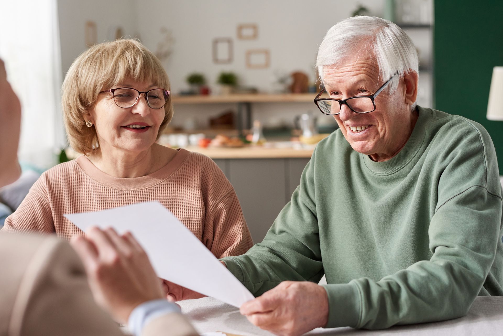 Two older adults sit at a table, smiling and holding a document while discussing senior living care options with another person whose back is to the camera.