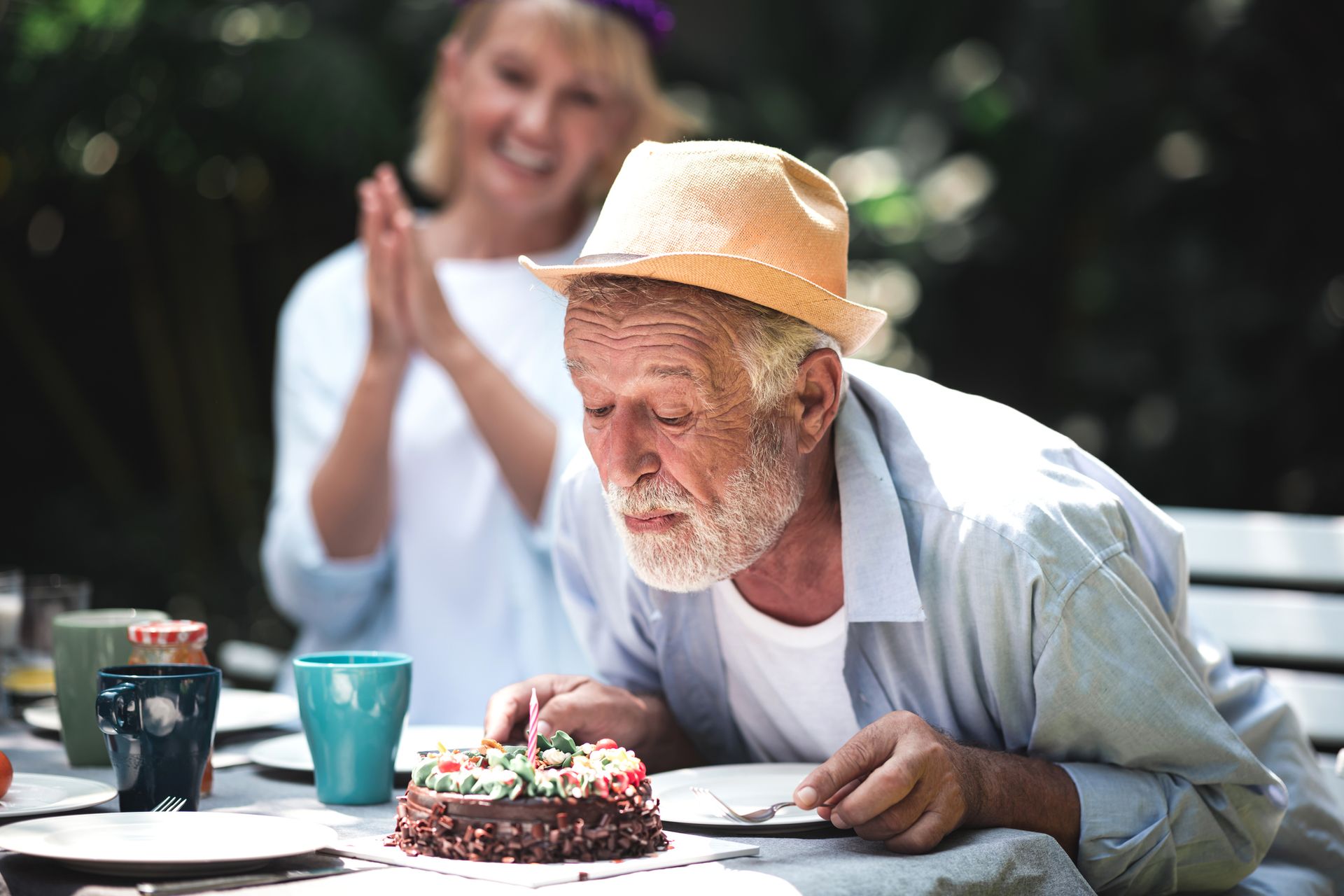 An elderly man wearing a hat blows out candles on a birthday cake while a smiling woman claps in the background at an outdoor table, celebrating joyful moments in senior living.