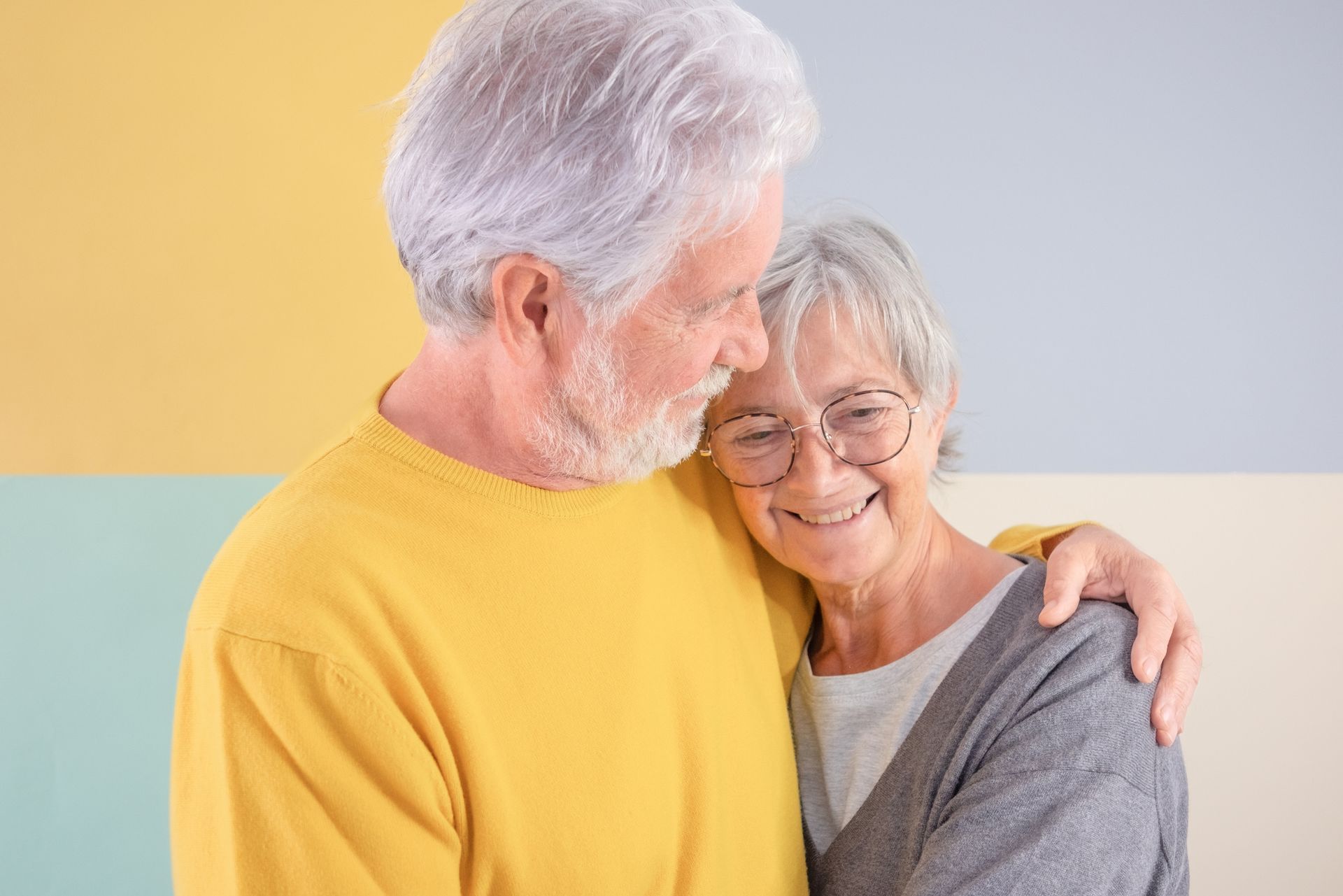 An older man in a yellow sweater hugs and smiles at an older woman in glasses and a gray cardigan, standing against a pastel-colored background—perfect for sharing questions to ask while touring senior living communities.
