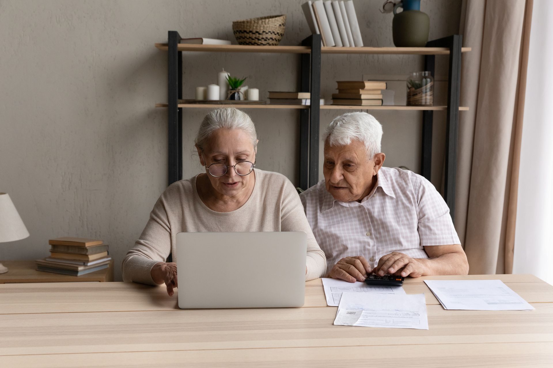 An elderly woman uses a laptop while an elderly man sits beside her with papers and a calculator, discussing senior living cost options in a cozy home office setting.