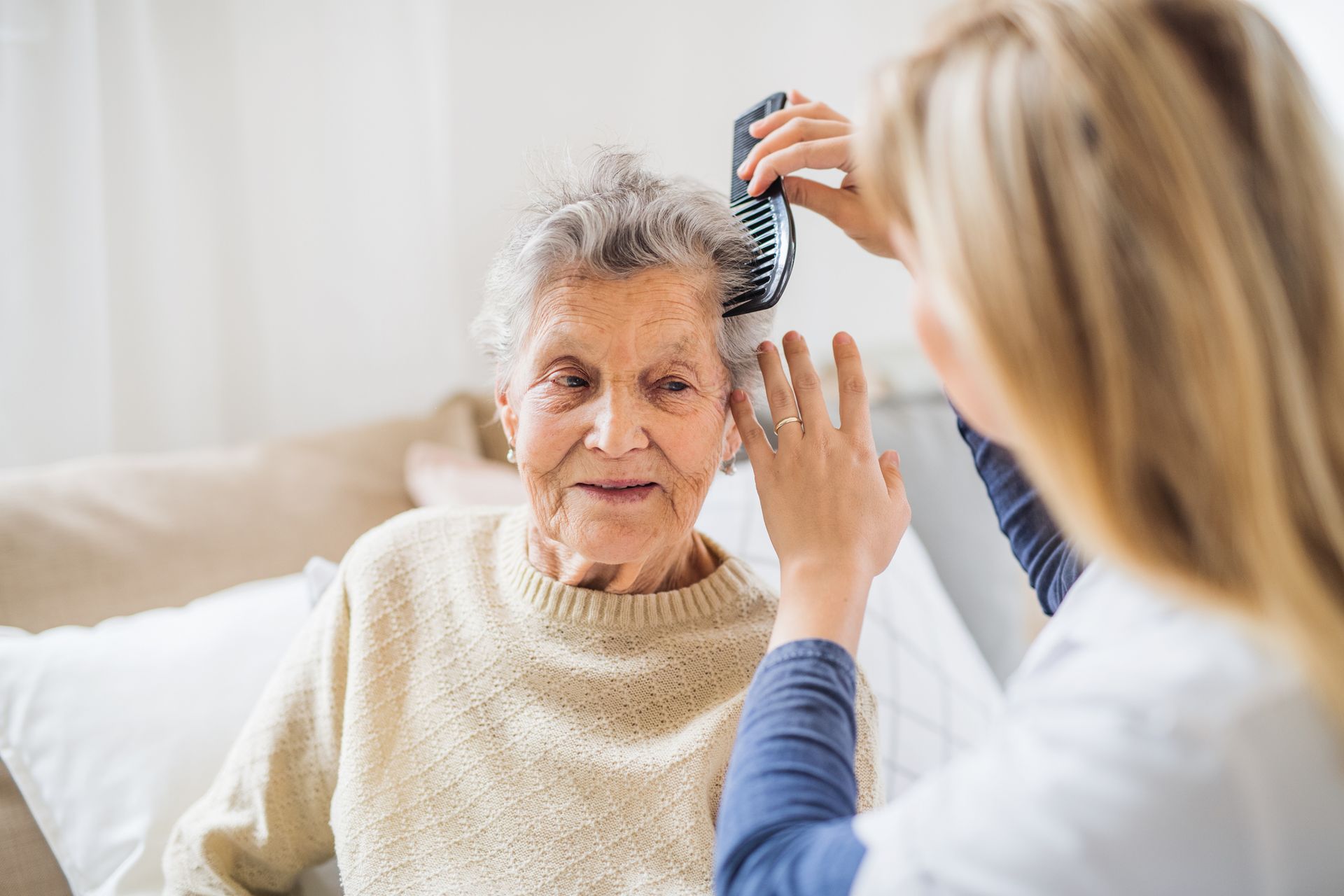 A caregiver provides personal care by gently brushing the hair of a senior woman sitting on a couch in a bright room.
