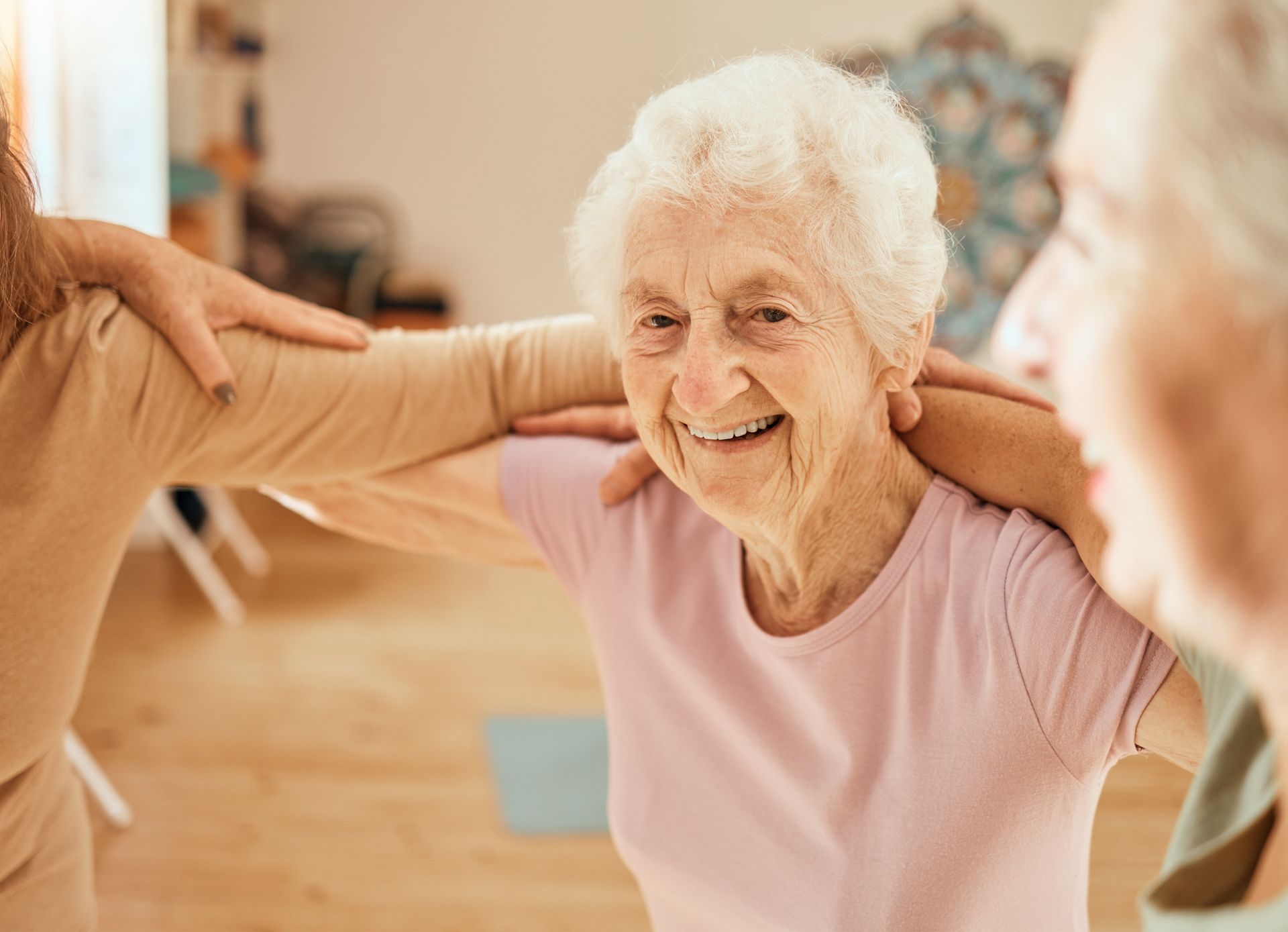 Elderly women standing in a circle with their hands on each other's shoulders, smiling and engaging in a group activity indoors, promoting senior socialization and enhancing senior wellbeing.