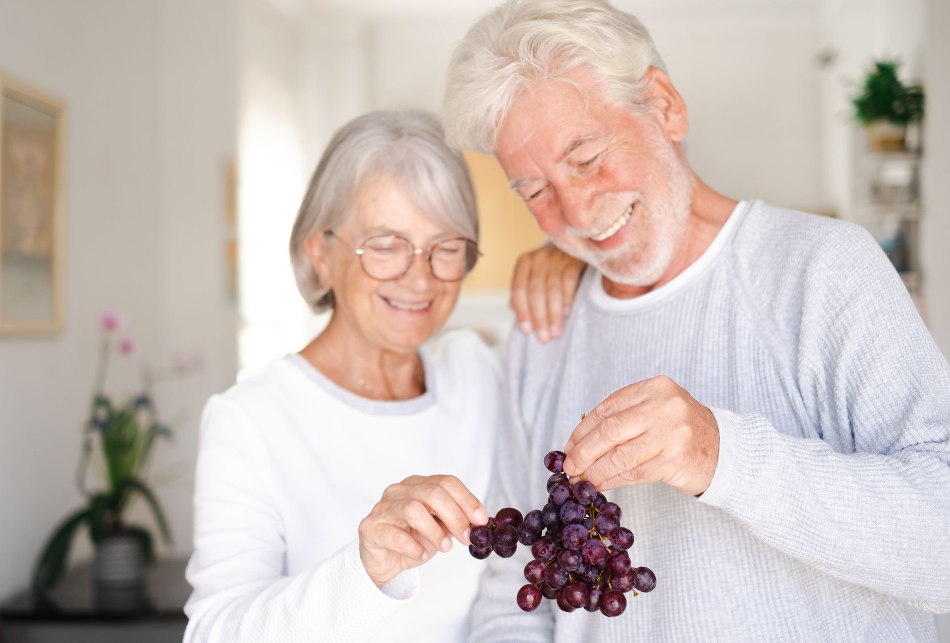 An older couple smiling together as they hold a bunch of grapes in a bright indoor setting, highlighting the importance of good nutrition for older adults.