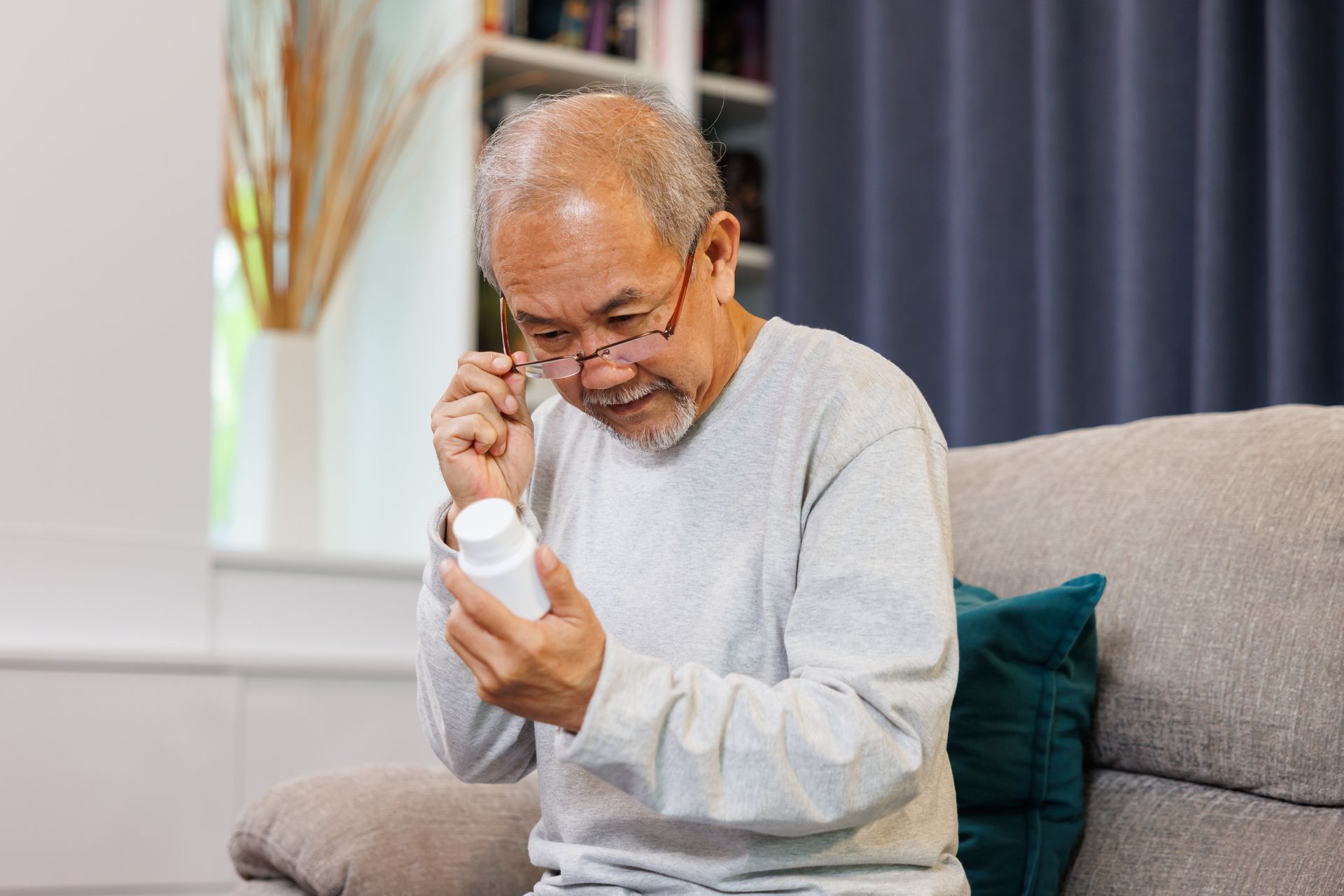 An older man sitting on a couch adjusts his glasses while closely reading the label on a white medicine bottle, taking care to avoid medication mix-ups—an important concern for seniors living at home.