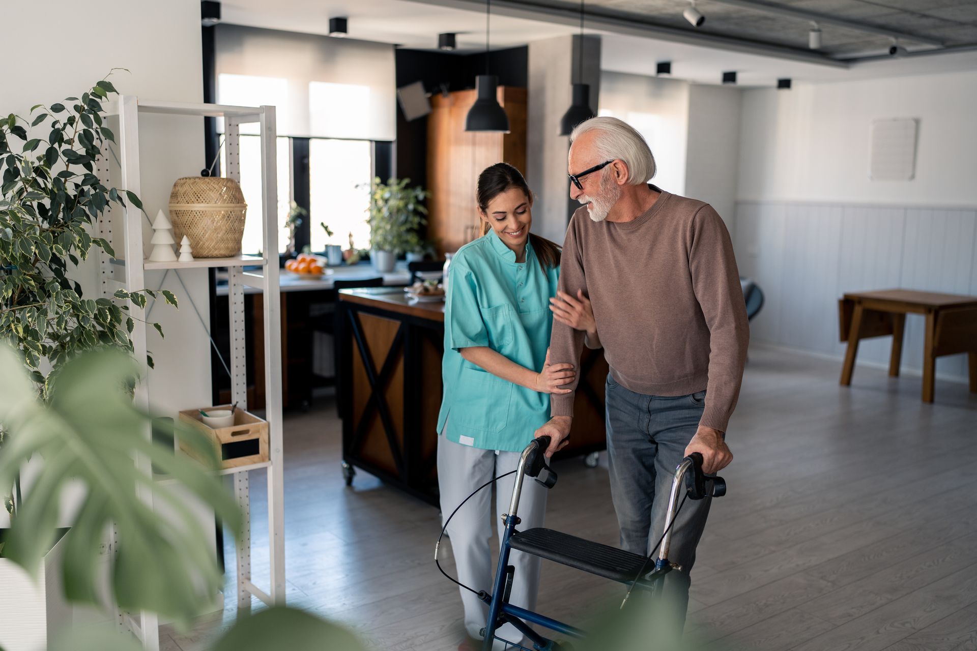 A caregiver assists an older man using a walker as they walk together in a modern, well-lit assisted living space, offering a glimpse of what to expect in supportive senior care environments.
