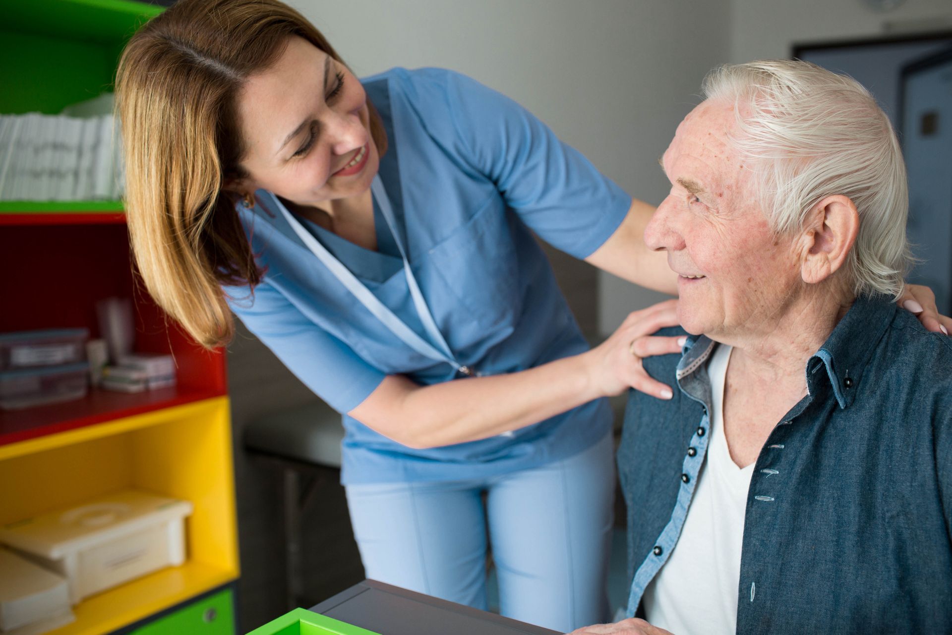 A female caregiver in blue scrubs smiles and touches the shoulder of an elderly man with white hair, who is sitting and looking at her in a brightly colored room, showing the compassion of comprehensive care on the road to recovery.