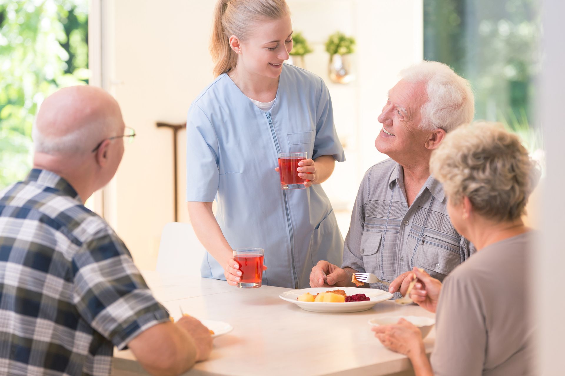 A caregiver serves drinks to three older adults seated at a dining table, with plates of food in front of them in a brightly lit assisted living community.