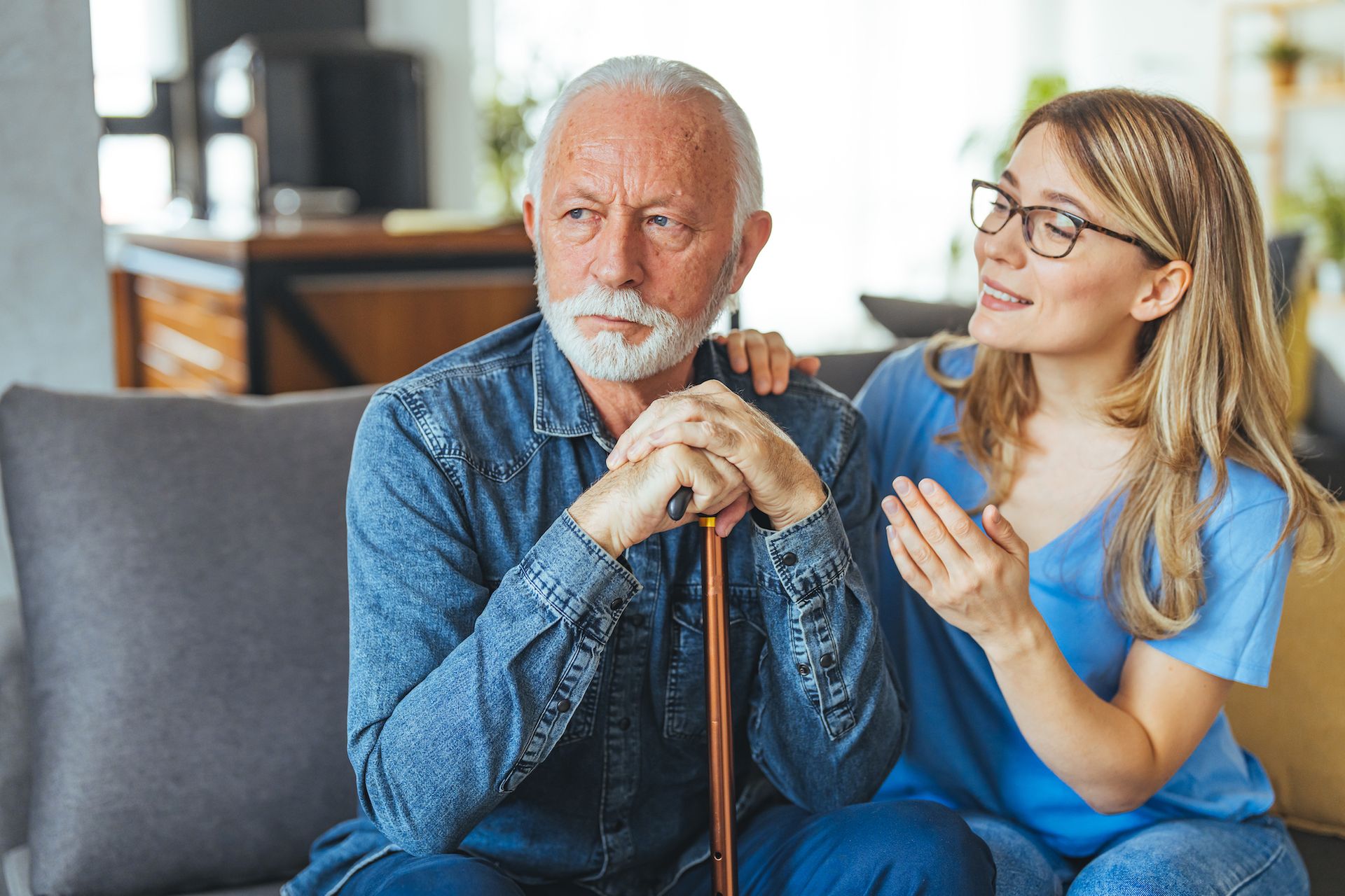 An older man with a cane sits on a couch looking away, while a younger woman beside him smiles and talks, resting her hand on his shoulder, showing the importance of support systems after traumatic brain injury.