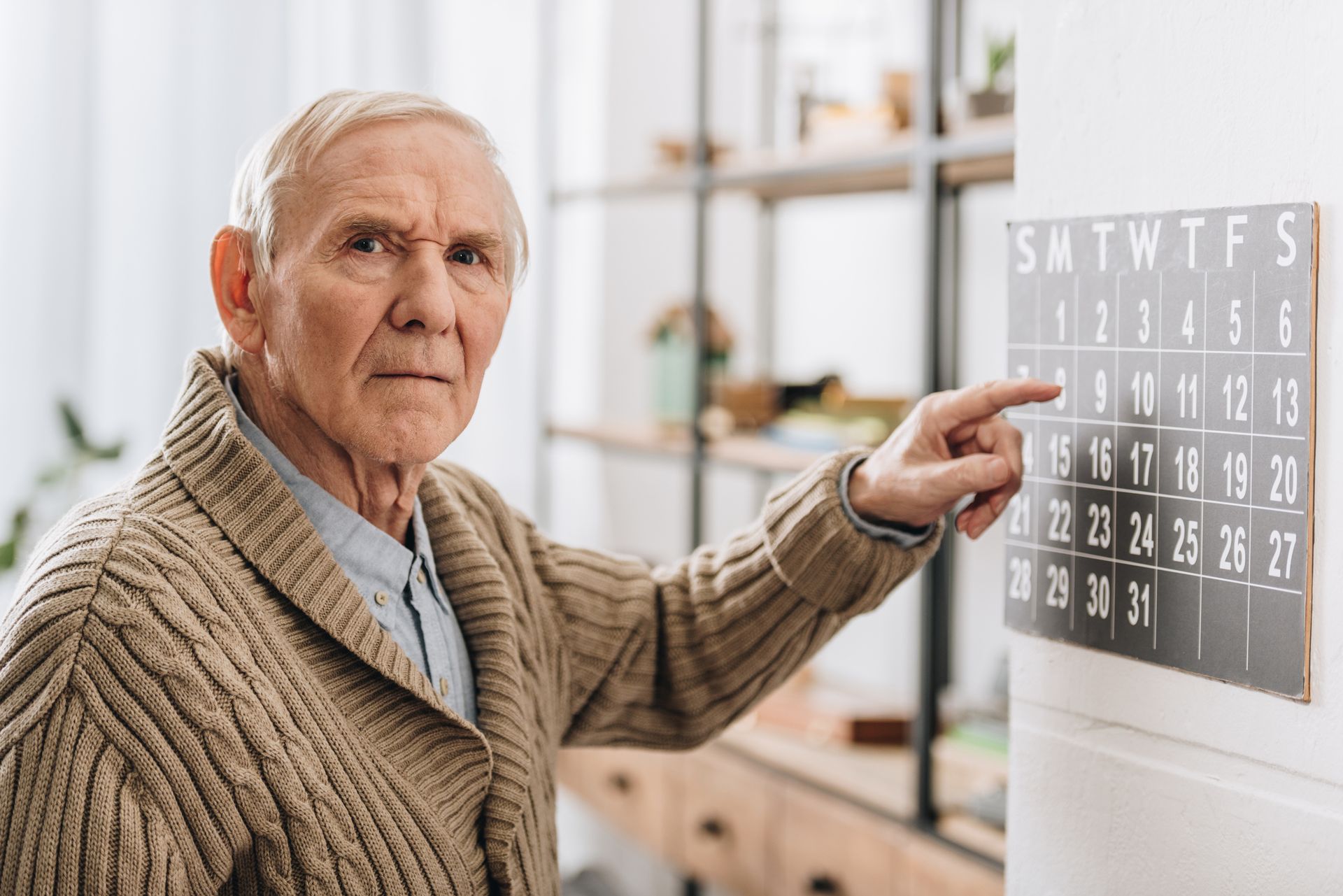 An elderly man in a beige sweater points at a date on a black wall calendar, highlighting how older adults manage daily routines amid cognitive changes, while looking directly at the camera in a well-lit room.