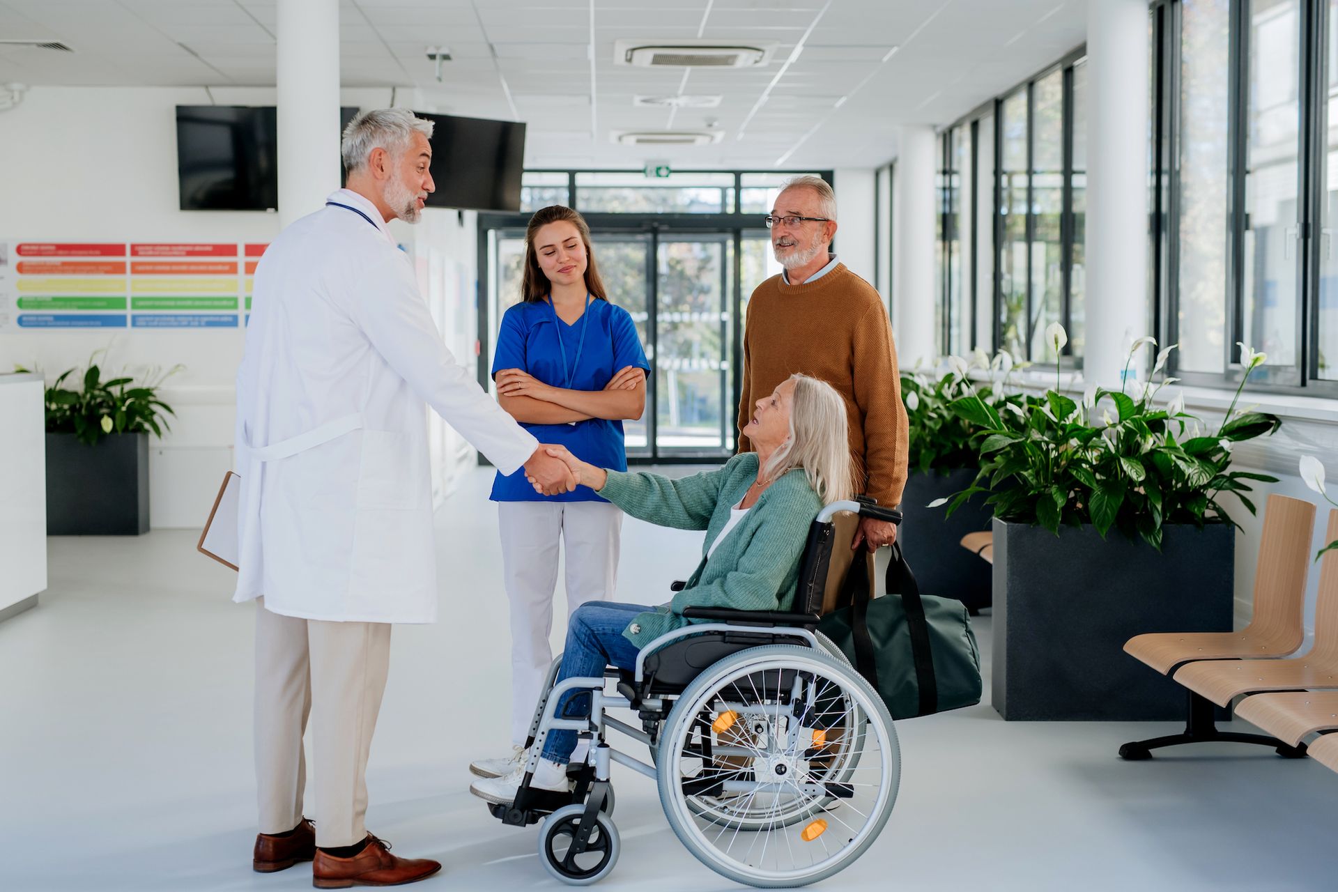 A doctor greets a woman in a wheelchair—who may have symptoms of a traumatic brain injury—accompanied by a nurse and an older man, in a bright hospital corridor filled with plants and seating.