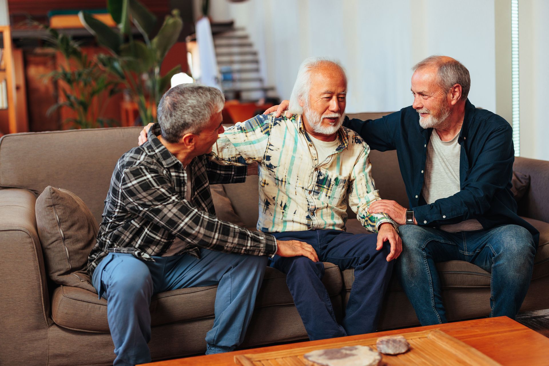Three older men sit together on a couch indoors, with two men comforting the man in the center, highlighting the importance of socialization for seniors and support to enhance senior well-being.