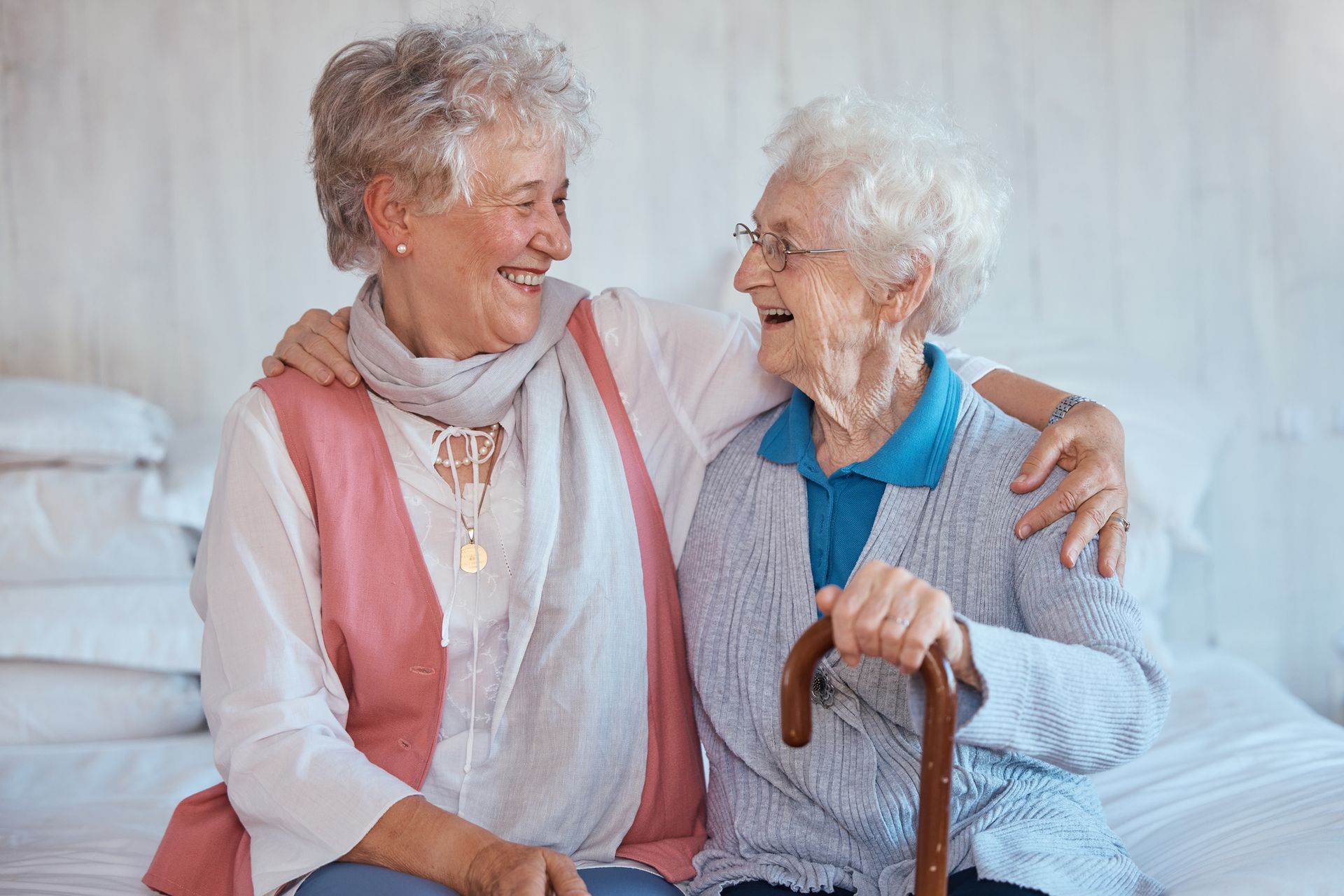 Two elderly women sit side by side on a bed in a shared living space, smiling at each other. One holds a cane, and they have their arms around each other in a close, affectionate manner.