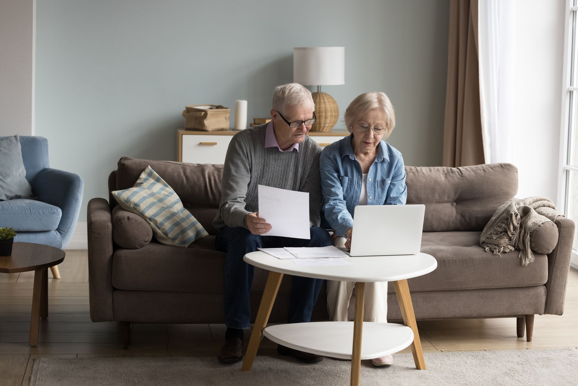 An older man and woman sit on a sofa looking at documents and a laptop, reviewing the cost of care at home in their living room.