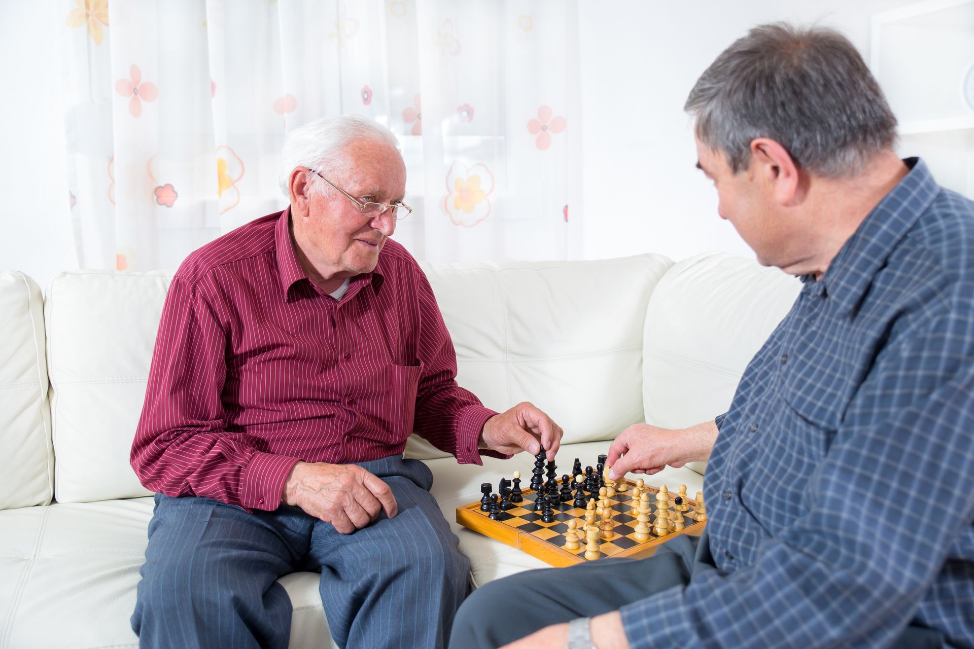 Two elderly men sit on a white couch and play a game of chess, embodying the daily life of memory care residents as one moves a piece while the other watches attentively.