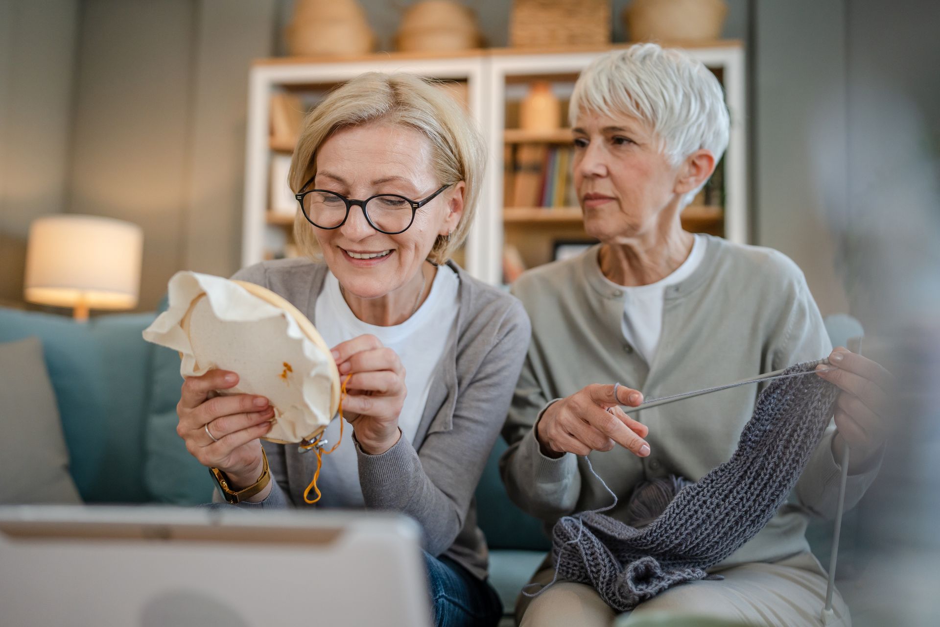 Two older women enjoy shared senior living, sitting on a couch; one embroiders with a smile as the other knits and looks at her. Shelves and home decor create a warm, inviting backdrop.