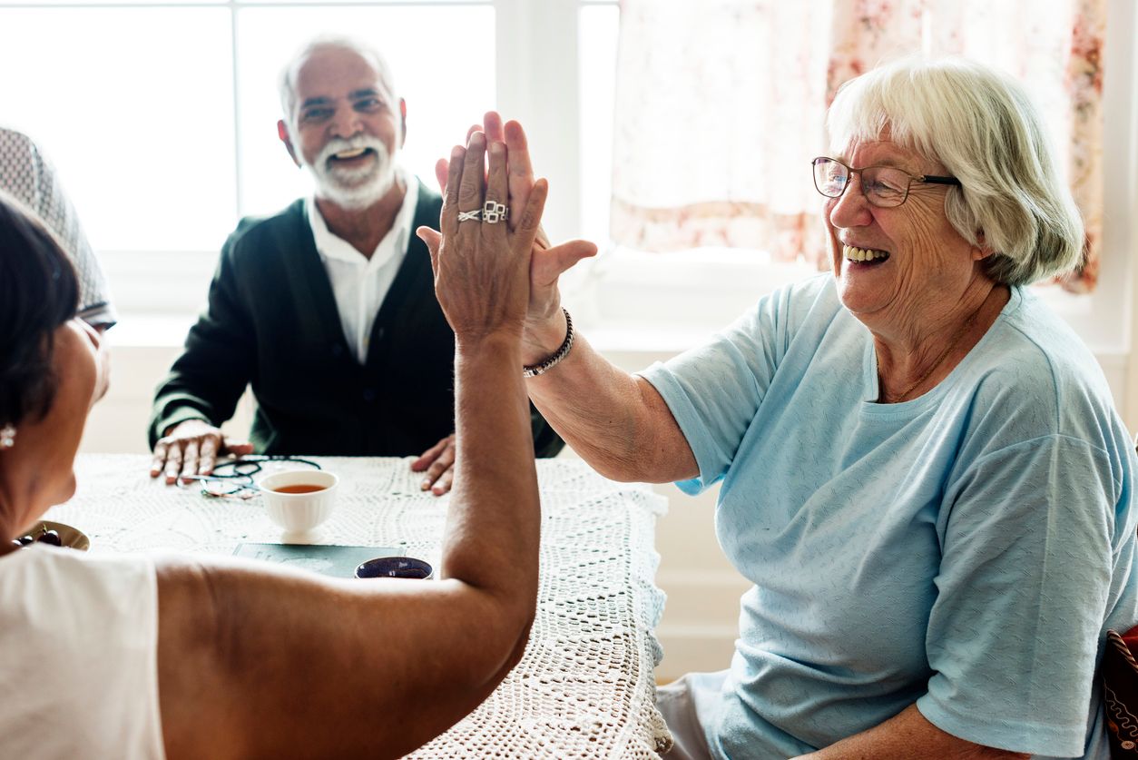 Two elderly residents high-five at a table while an elderly man smiles in the background. Drinks and a pair of glasses sit on the table, capturing a joyful moment in their shared living environment.