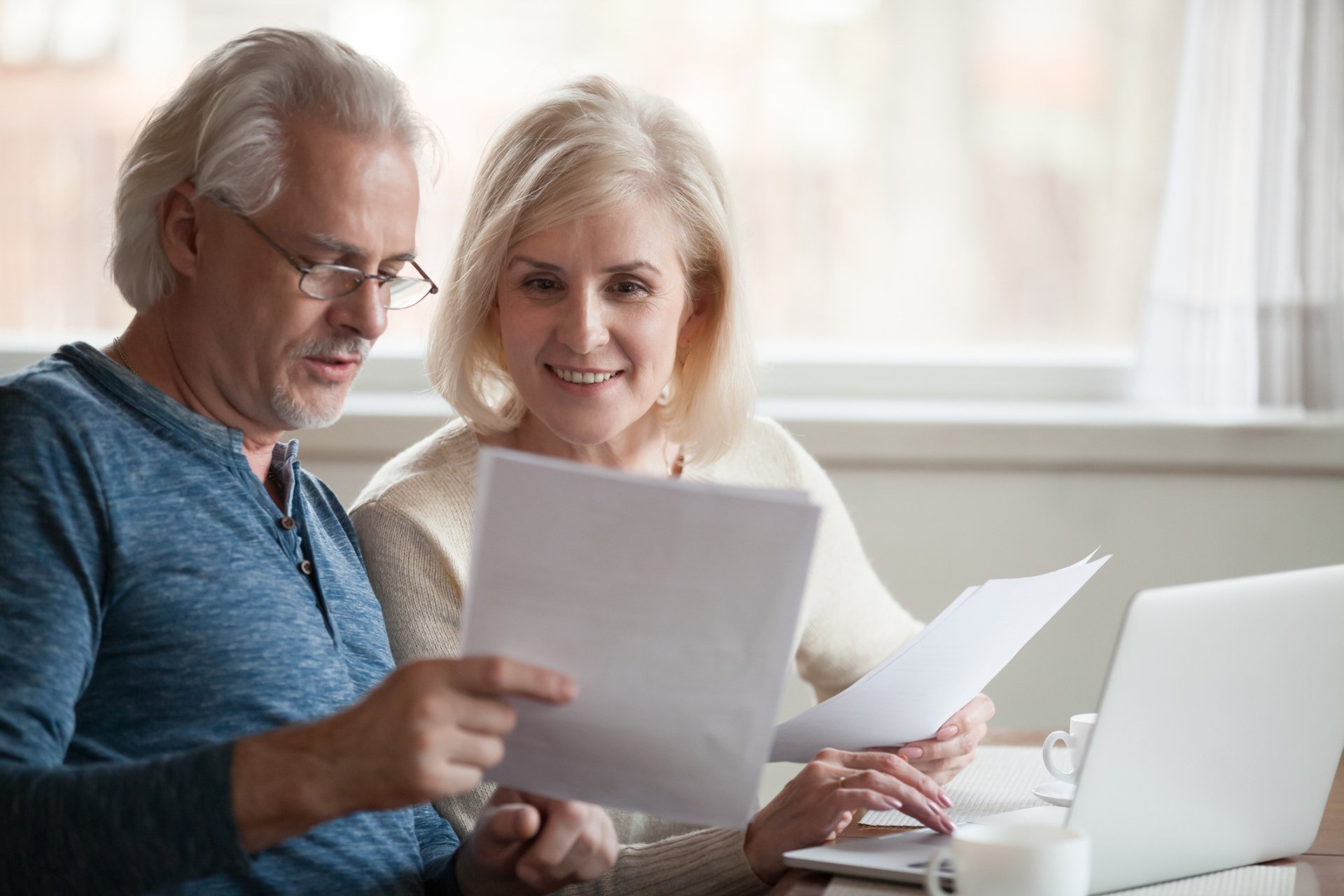 An older man and woman sit at a table reviewing documents related to senior housing, with a laptop and coffee cups nearby.