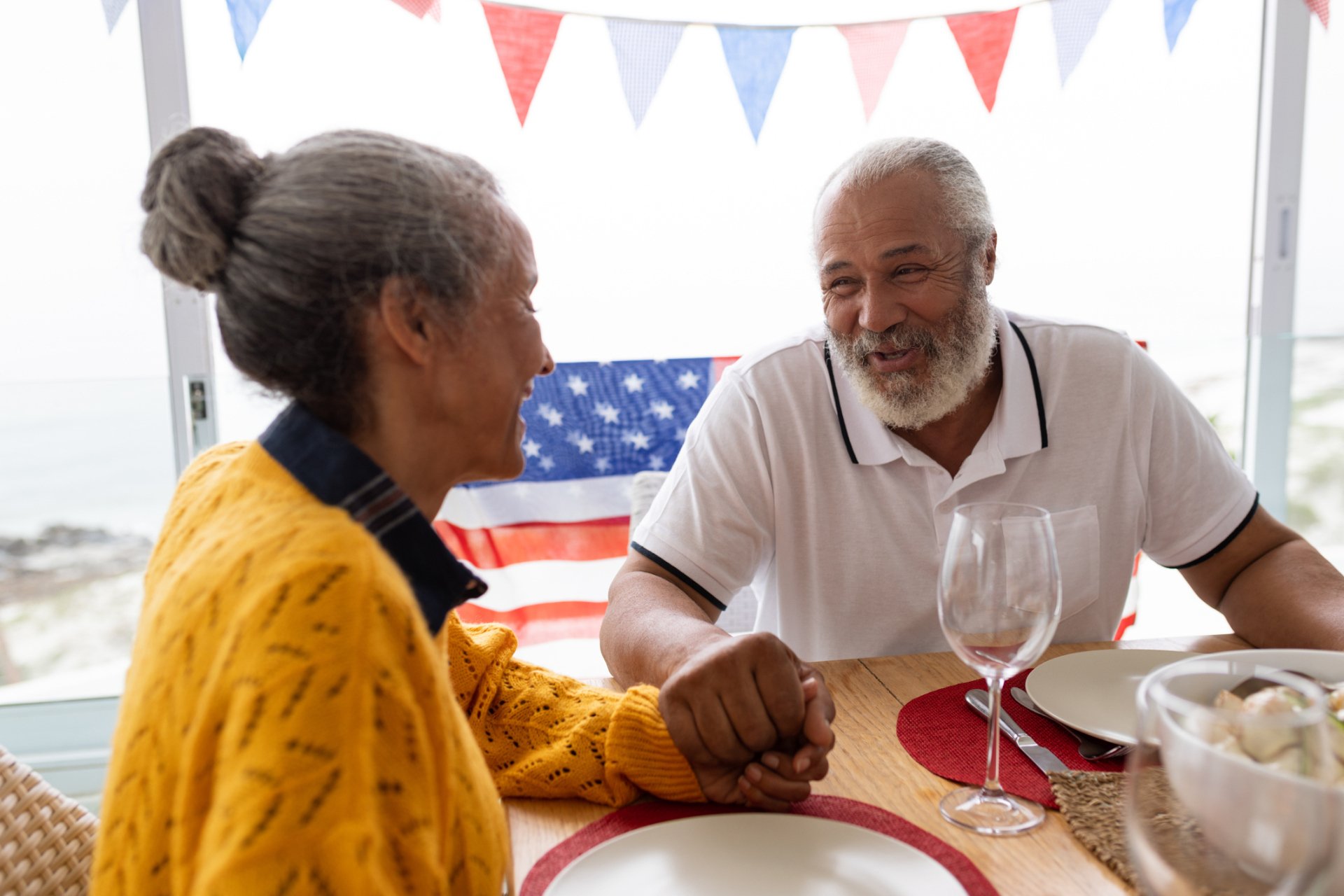 Two older adults sit at a table holding hands, smiling, with an American flag and festive bunting in the background, enjoying the warmth of senior living and honoring traditions.