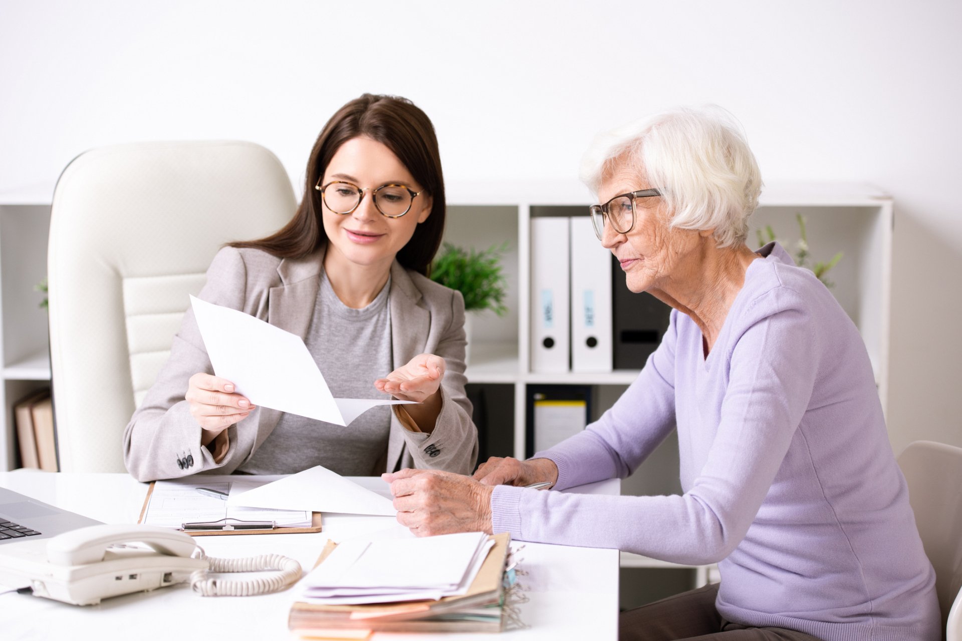 A younger woman shows a document to an older woman at a desk in an office setting, discussing insurance options, with papers, a pen, and a phone visible.