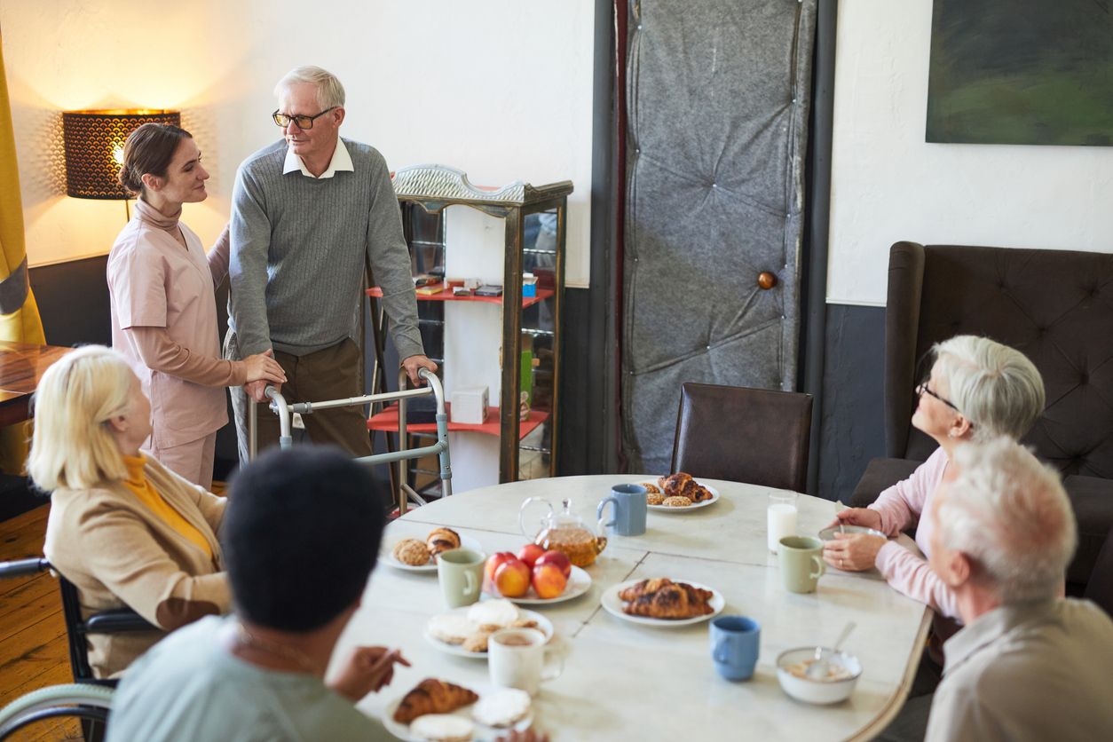 A caregiver assists an elderly man with a walker, while four other seniors with dementia sit at a round table enjoying breakfast in a communal dining area, highlighting compassionate dementia care in a shared living environment.