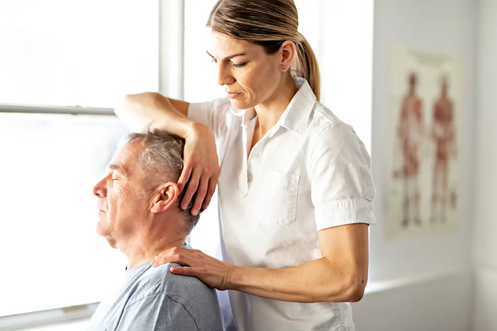 Assured-img-TBI3 A healthcare professional performs a neck and shoulder assessment on an older man seated near a window in a medical office.