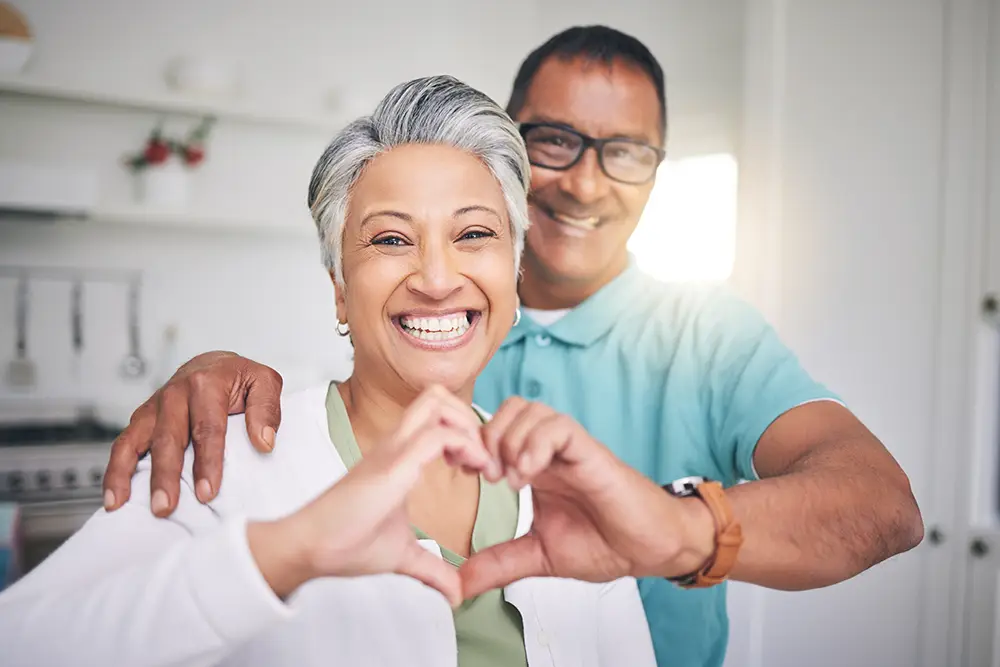 Smiling older couple stands in a bright kitchen; the woman forms a heart shape with her hands while the man stands behind her with an arm around her shoulder.