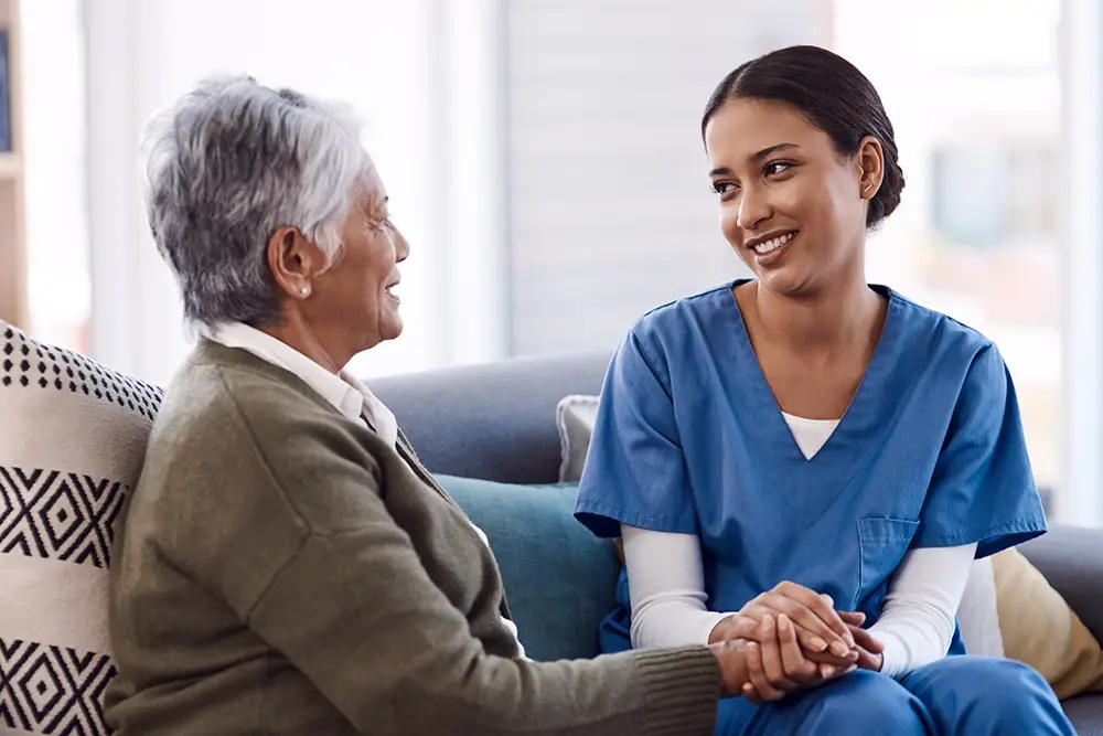 Assured-img5 A nurse in blue scrubs sits on a couch, holding hands and smiling with an older woman who is also smiling.