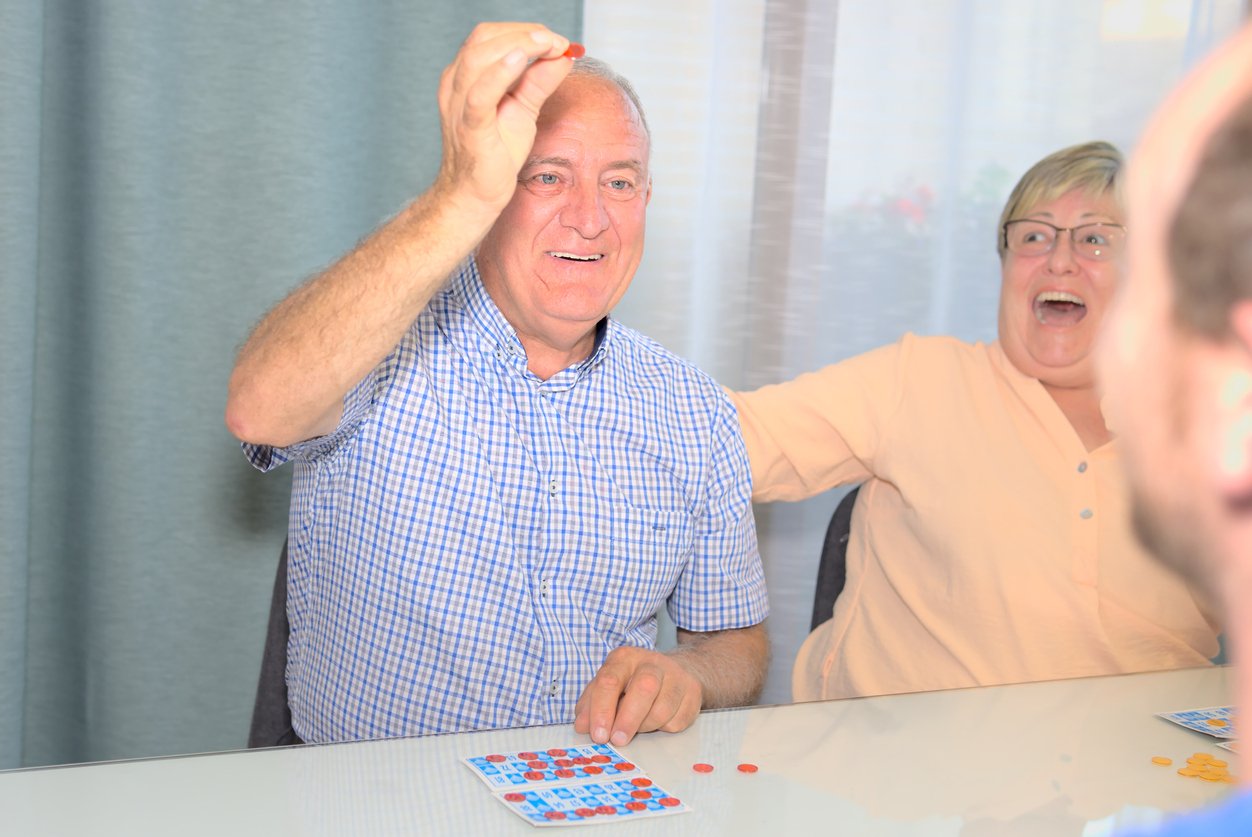 Residents enjoy daily activities as an older man smiles and raises a bingo chip while a woman beside him laughs; both are sitting at a table with bingo cards and red chips in this vibrant senior living community.