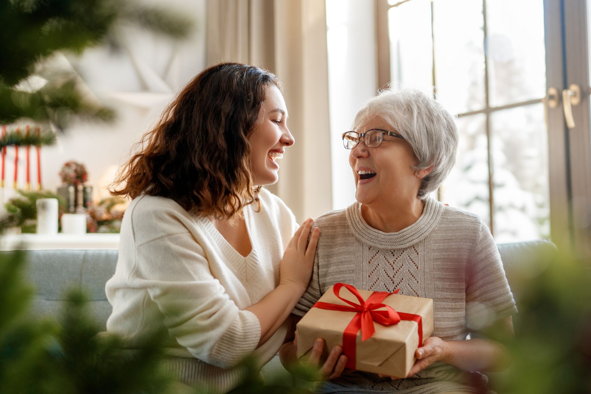 Two women sit on a couch, smiling at each other. The older woman, enjoying celebrations for seniors, holds a gift box with a red ribbon. A decorated tree and festive holiday traditions are visible in the background.