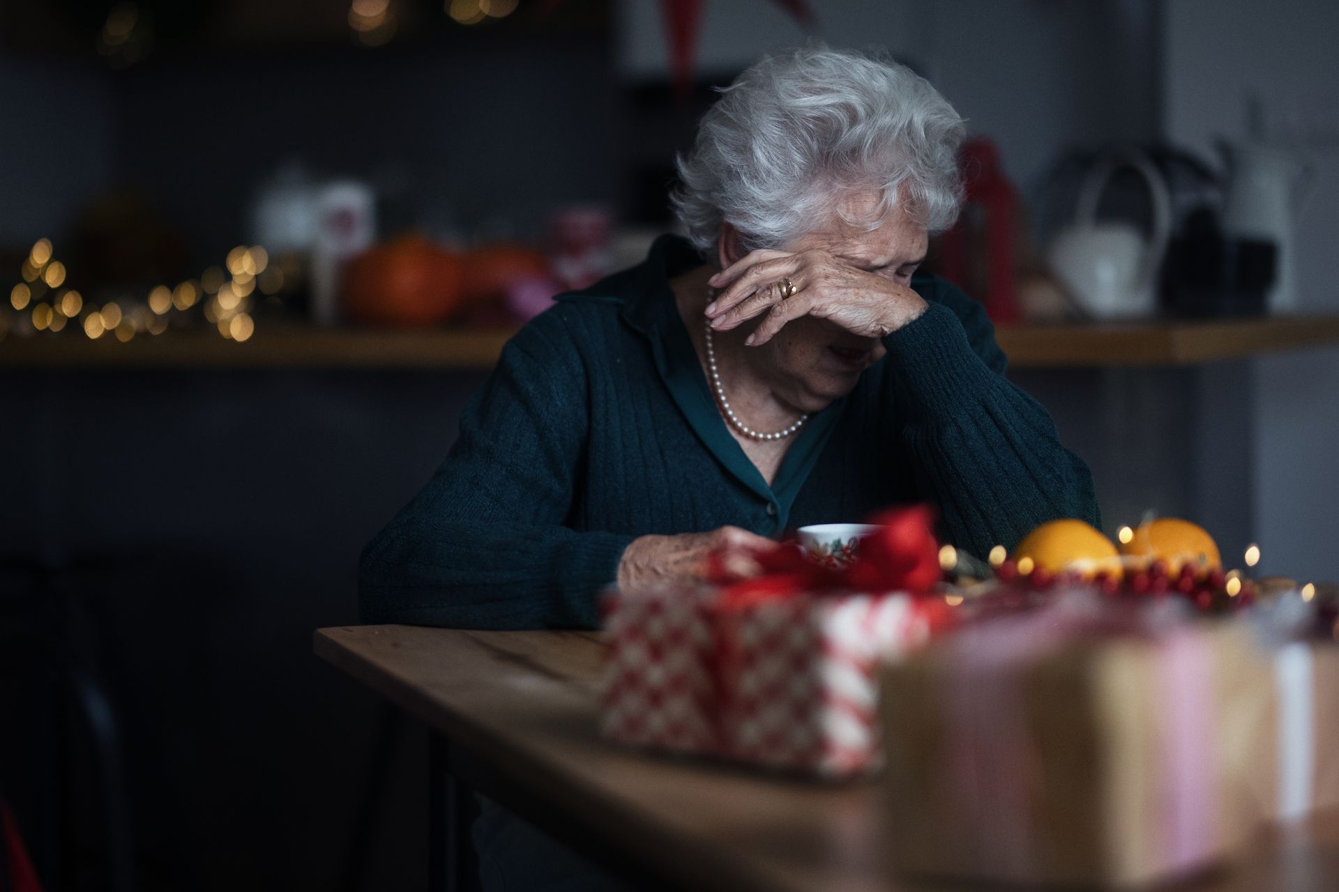 An elderly person sits at a table with wrapped gifts, holding a cup and covering their face, appearing emotional or upset in a dimly lit room—a poignant moment reflecting holidays and grief.