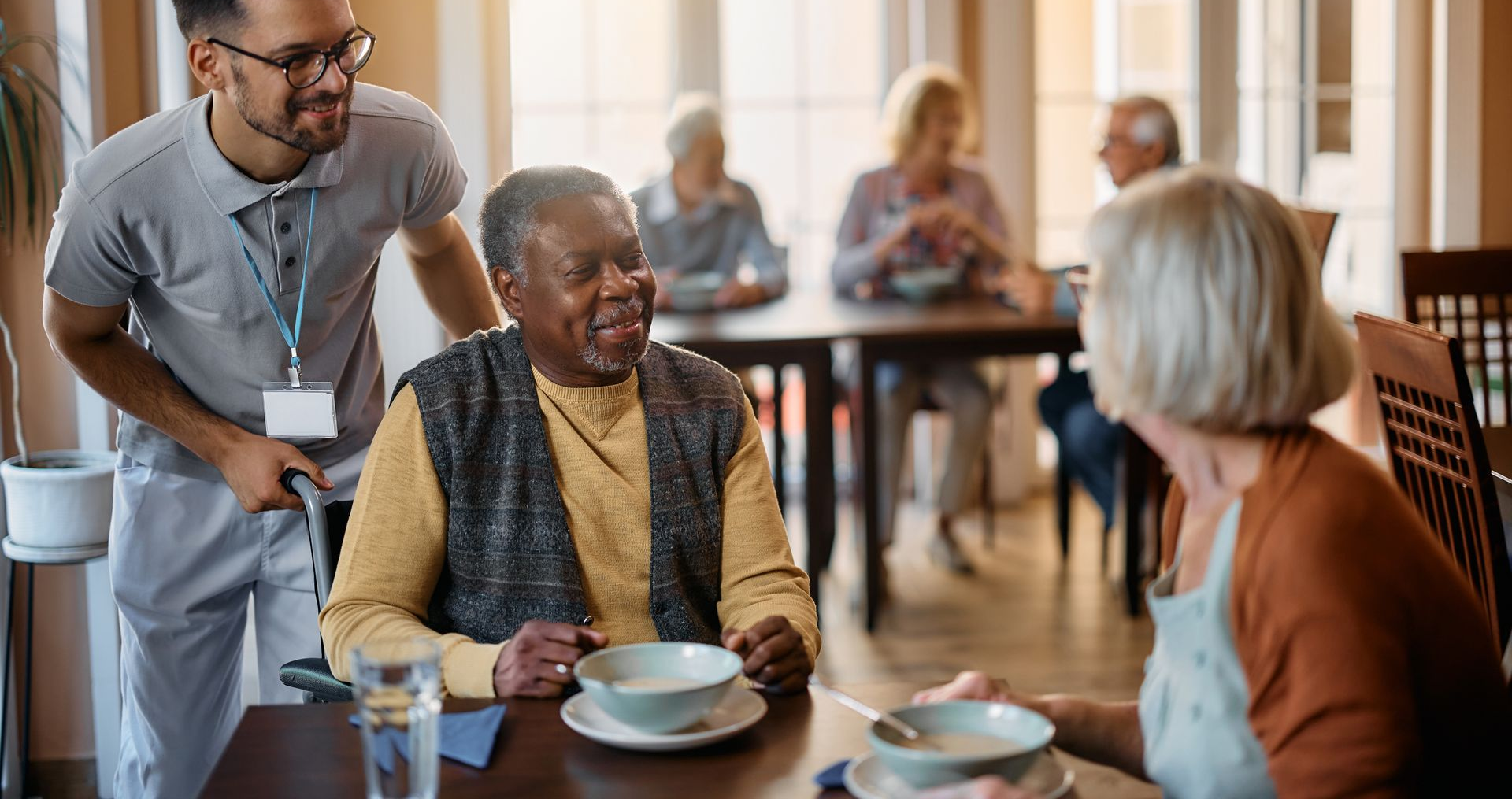 A caregiver assists a man in a wheelchair at a dining table with another woman in a communal setting, where others converse—showcasing loved ones enjoying quality time assisted living together.