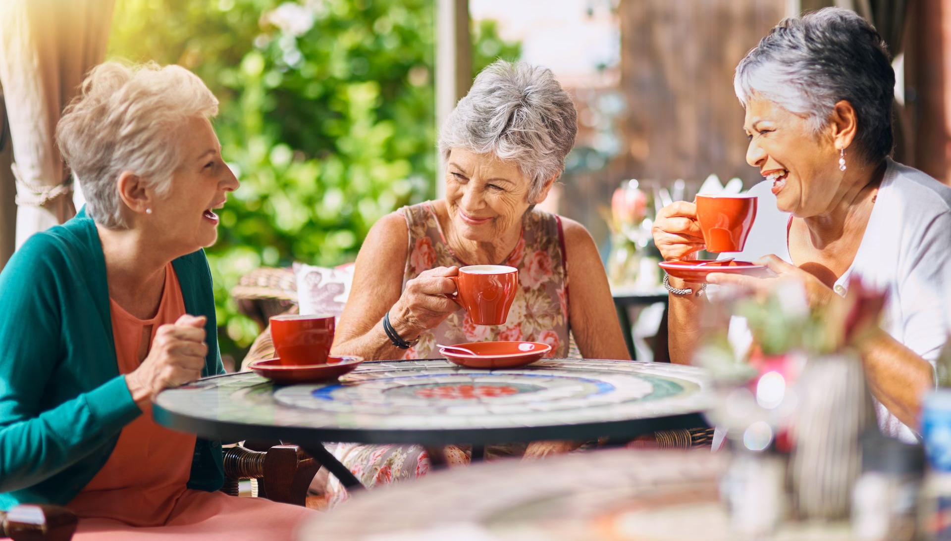 Three seniors sit at a round outdoor table, smiling and talking while holding red cups, enjoying the greenery as they share wisdom and life stories together.