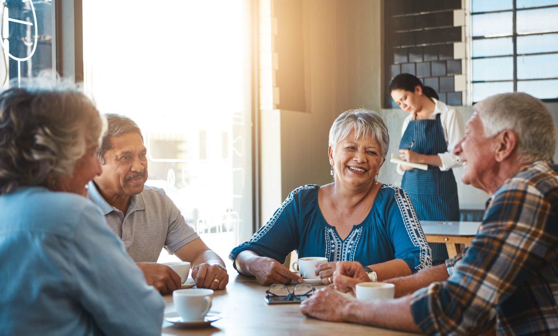 Four older adults sit at a café table, smiling and sharing memories over coffee, as a waitress works in the background.