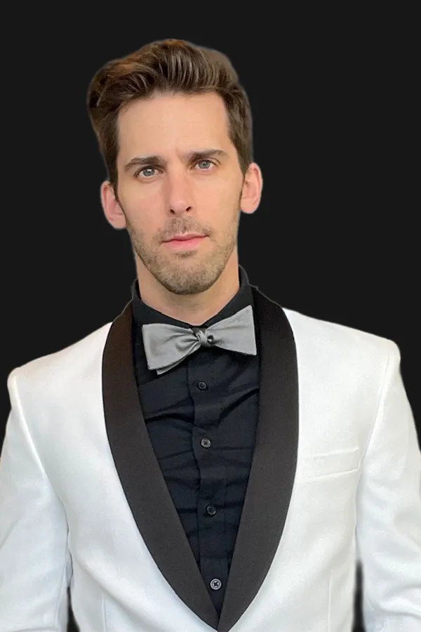 A man wearing a white tuxedo jacket with black lapels, a black shirt, and a silver bow tie stands against a plain black background.