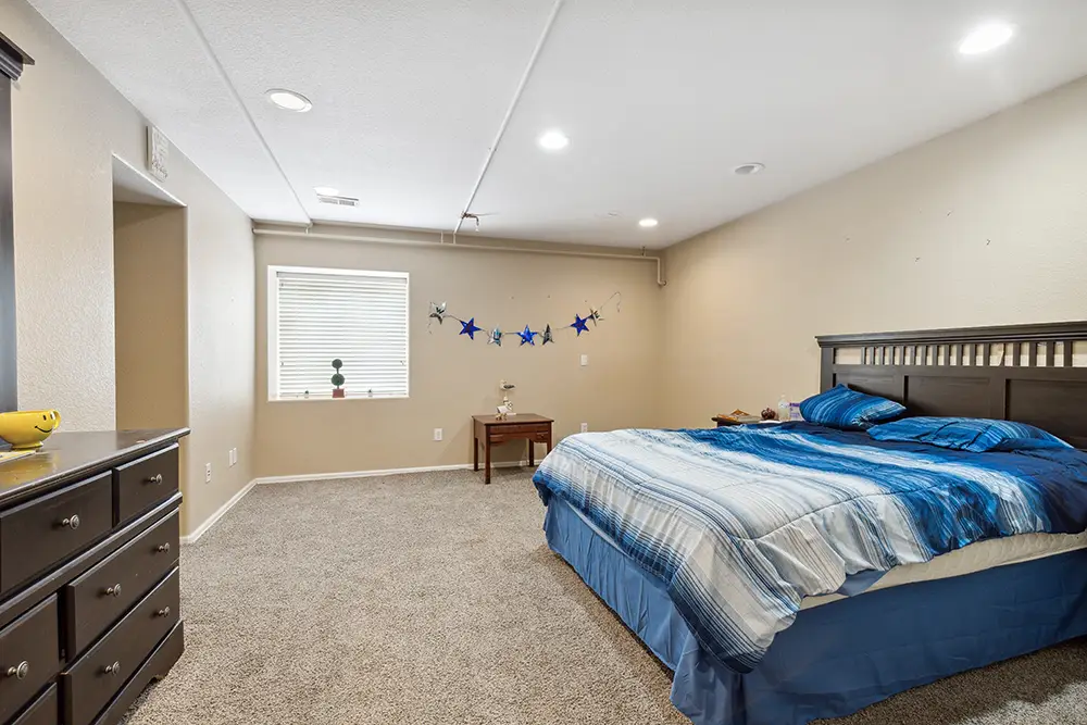 A bedroom with a large bed, blue bedding, carpeted floor, dresser, small table, and a window with a star garland on the wall.