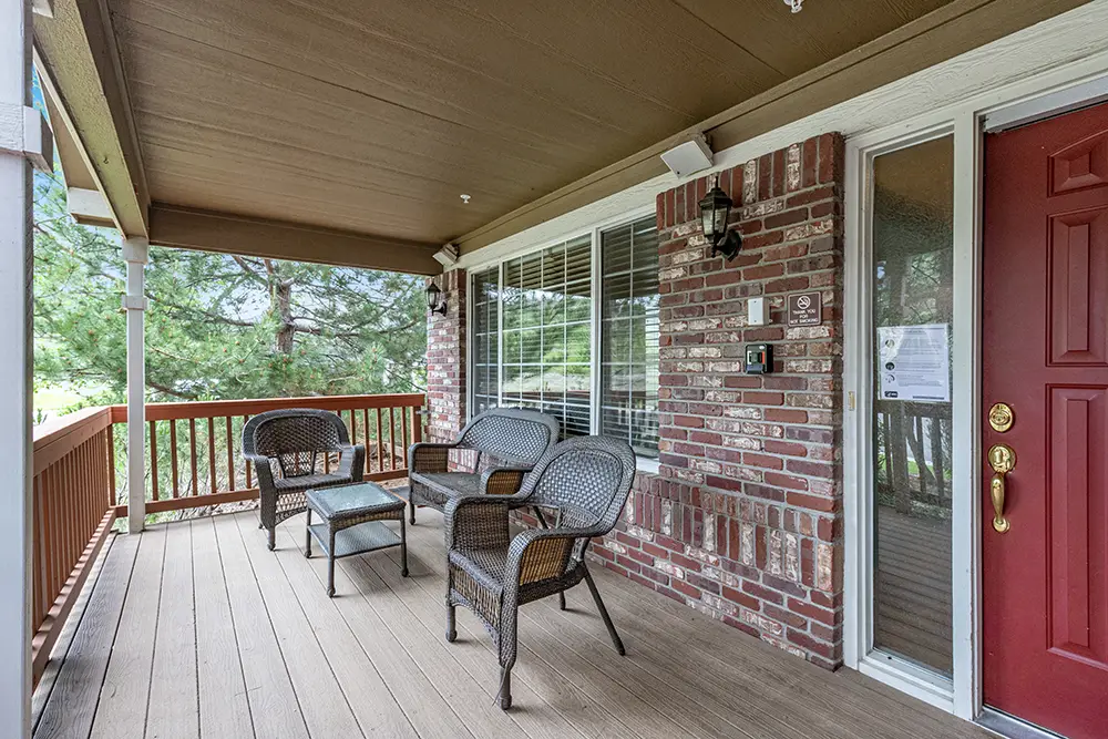 Covered porch with three wicker chairs, a glass-topped table, brick exterior walls, large windows, and a red front door. Trees and greenery are visible in the background.