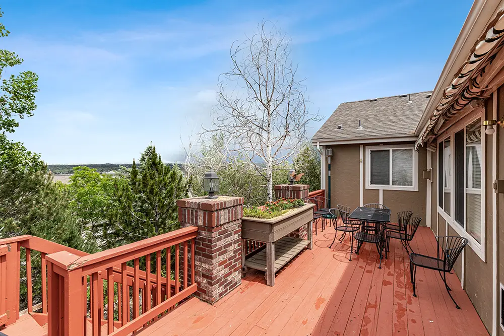 Spacious red wooden deck with outdoor seating, potted tree, brick columns, and view of greenery under a clear blue sky.