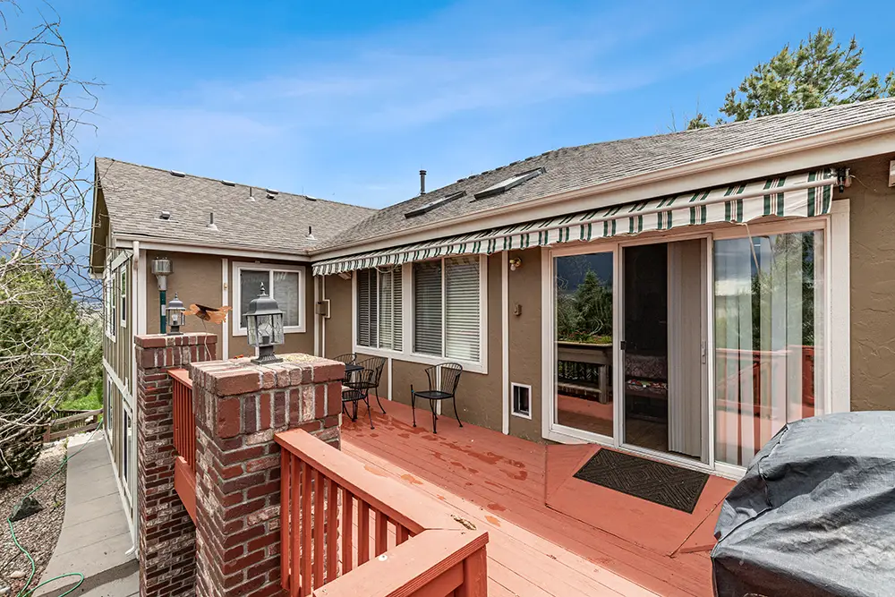 Wooden deck attached to tan house with sliding glass doors, striped awning, red railing, outdoor seating, and brick posts, set under a blue sky.