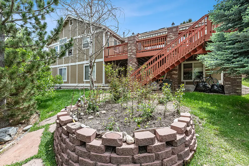 A raised circular garden bed made of bricks is in the yard of a two-story house with a wood deck and exterior stairs.