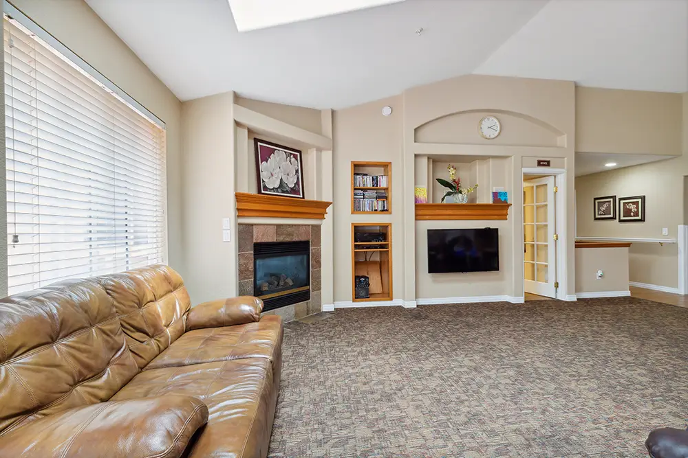 Living room with brown leather sofa, wall-mounted TV, built-in shelves, fireplace, floral paintings, carpeted floor, and large window with blinds.