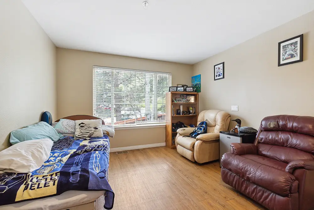 A tidy bedroom with a single bed, two recliner chairs, a bookshelf, framed photos, and a window with blinds letting in natural light.