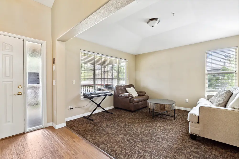 Living room with a brown armchair, beige sofa, round coffee table, keyboard stand, carpeted floor, large windows, and neutral walls.