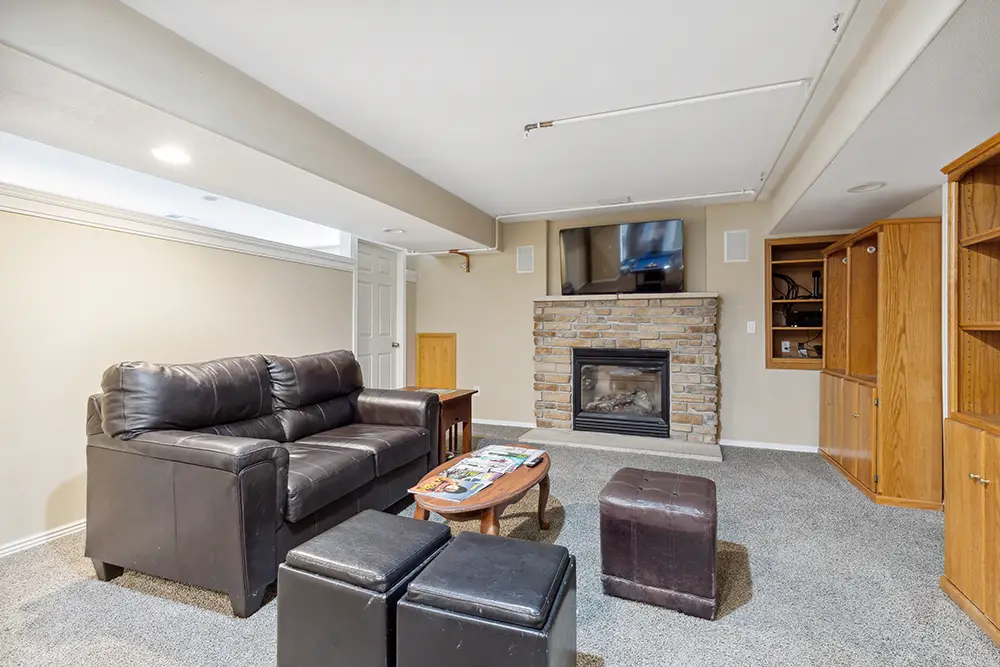 Basement living room with a brown leather couch, ottomans, a wooden coffee table, brick fireplace, wall-mounted TV, and built-in wooden shelves on carpeted flooring.
