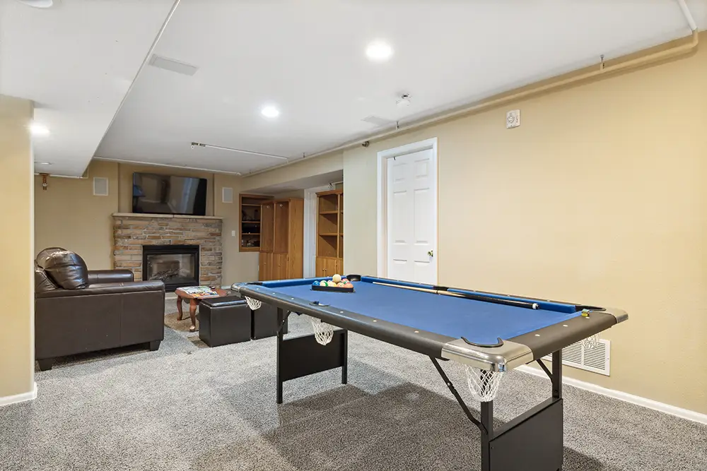 A basement room with a blue pool table, a brown couch and chair, a fireplace with a TV above it, built-in shelves, and carpeted floor.