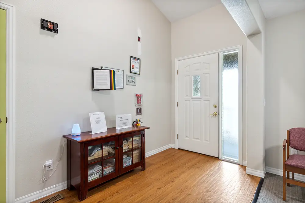 A small waiting area with a wooden cabinet, informational papers, and chairs by a white front door with frosted glass panels.
