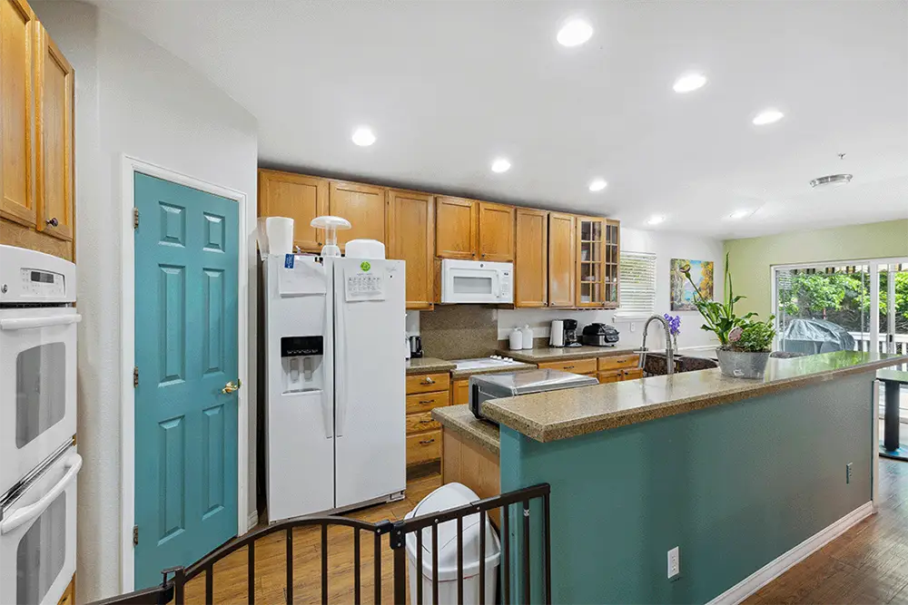 Modern kitchen with wooden cabinets, teal pantry door, white appliances, brown countertop island, and recessed ceiling lights; sliding door opens to a patio with outdoor greenery visible.