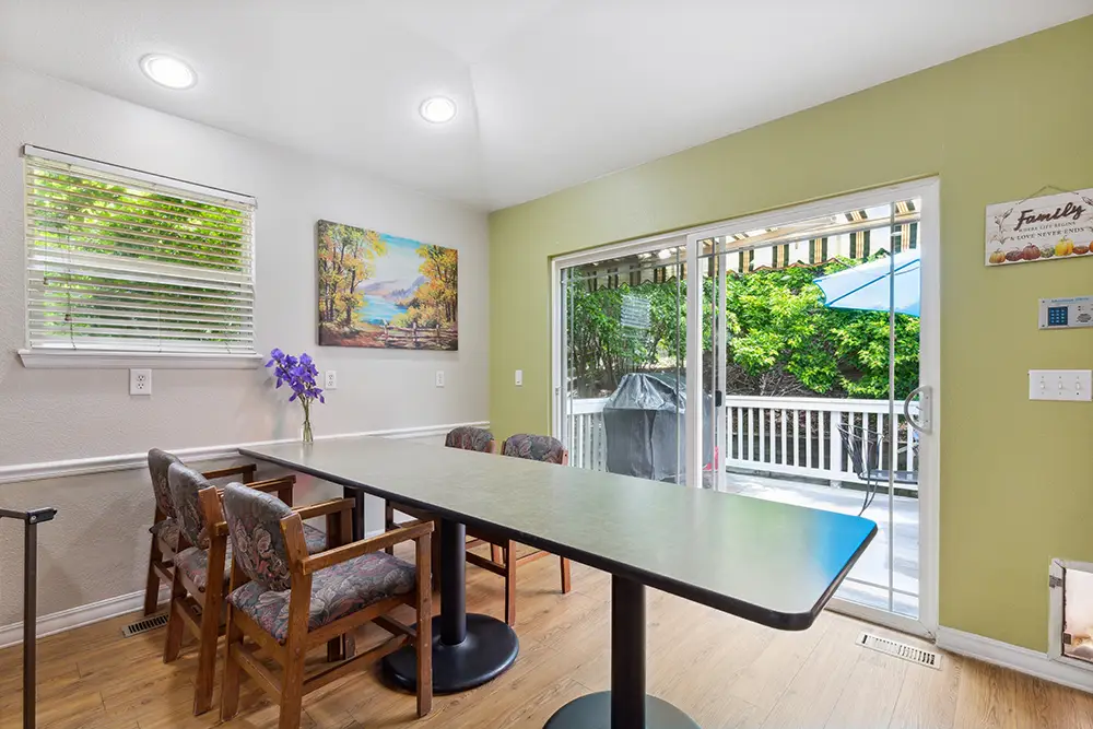 Dining area with a long table and four chairs beside sliding glass doors leading to a deck with a grill and patio umbrella; window and nature artwork on walls, wood floor.