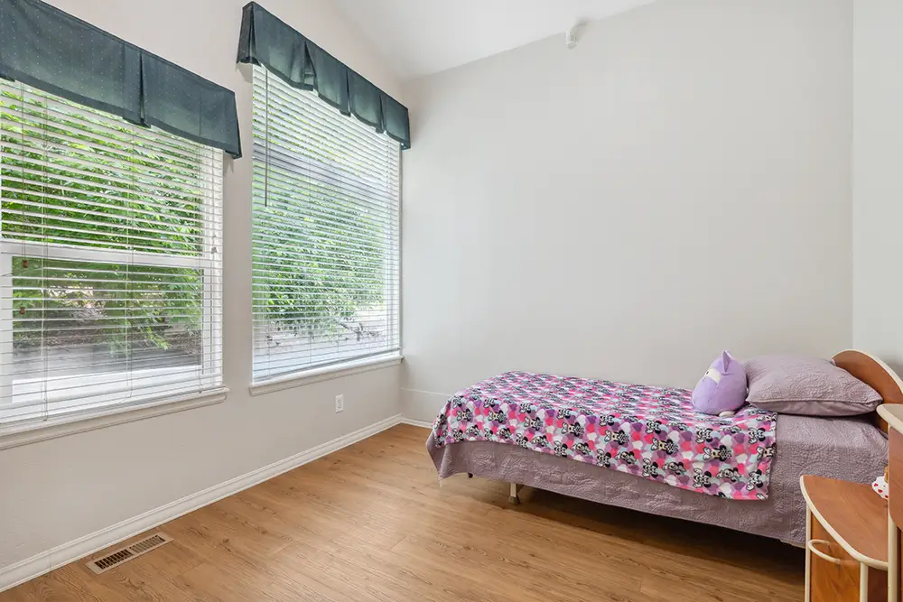 Small bedroom with a single bed, floral blanket, pillows, hardwood floor, large windows with blinds, and green valances.